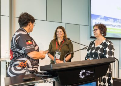 Kellie Kerin passing the Message Stick - a col­lec­tive mes­sage of Cul­tur­al Safe­ty, craft­ed by the group, was then tak­en for­ward and passed on to the wider conference - to Linda Kensington and Dallas McKeown.